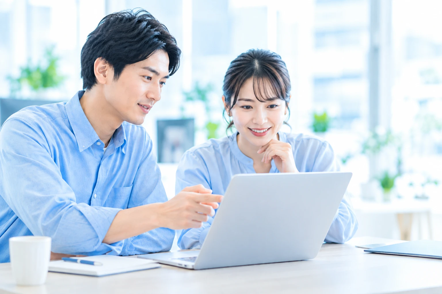 明るいオフィスで、2人の会社員がノートパソコンの画面を一緒に見ながら相談している写真風イメージ